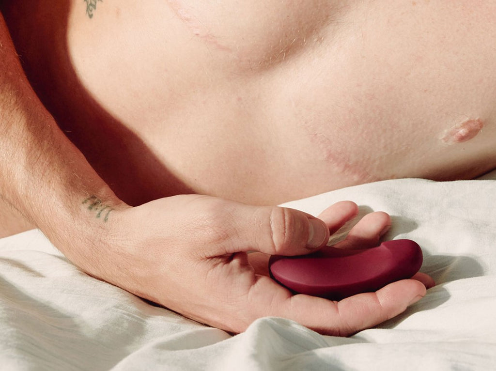 A person with top surgery lays on a bed while holding a Dame vibrating toy